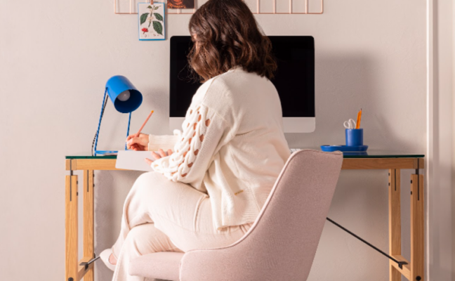 decoração home office pequeno com mulher sentada em uma cadeira de escritório fazendo anotações em um caderno em cima de uma escrivaninha, além de luminária de mesa, monitor e um quadro para pendurar anotações.