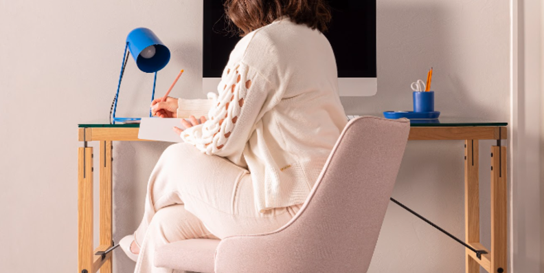 decoração home office pequeno com mulher sentada em uma cadeira de escritório fazendo anotações em um caderno em cima de uma escrivaninha, além de luminária de mesa, monitor e um quadro para pendurar anotações.