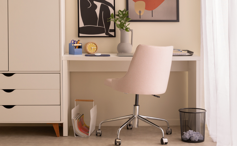 home office pequeno no canto do quarto com escrivaninha, cadeira de escritório com rodinhas, lixeira, quadros decorativos e um guarda-roupas branco ao lado.