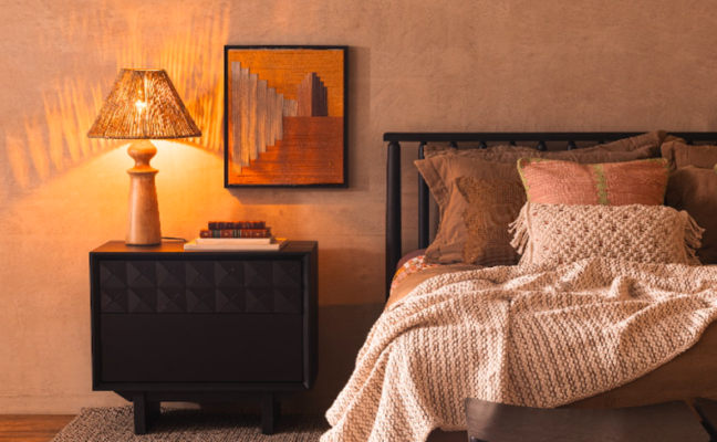 iluminação indireta quarto com mesa de cabeceira, abajur com luz amarela, cama, cabeceira, edredom e diversos travesseiros e almofadas, além de um quadro decorativo.