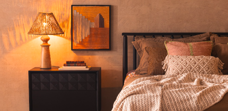 iluminação indireta quarto com mesa de cabeceira, abajur com luz amarela, cama, cabeceira, edredom e diversos travesseiros e almofadas, além de um quadro decorativo.