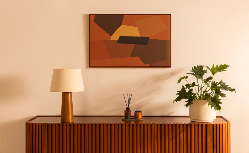luminária de mesa em um buffet na sala de estar, além de vaso com planta, aromatizador de ambiente e um quadro decorativo.