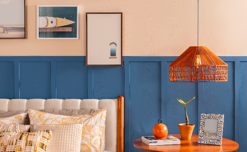 pendente iluminação indireta no quarto de casal com pendente acima de uma mesa de cabeceira, cama com cabeceira branca, diversas almofadas, uma parede pintada metade de azul e diversos quadros decorativos.