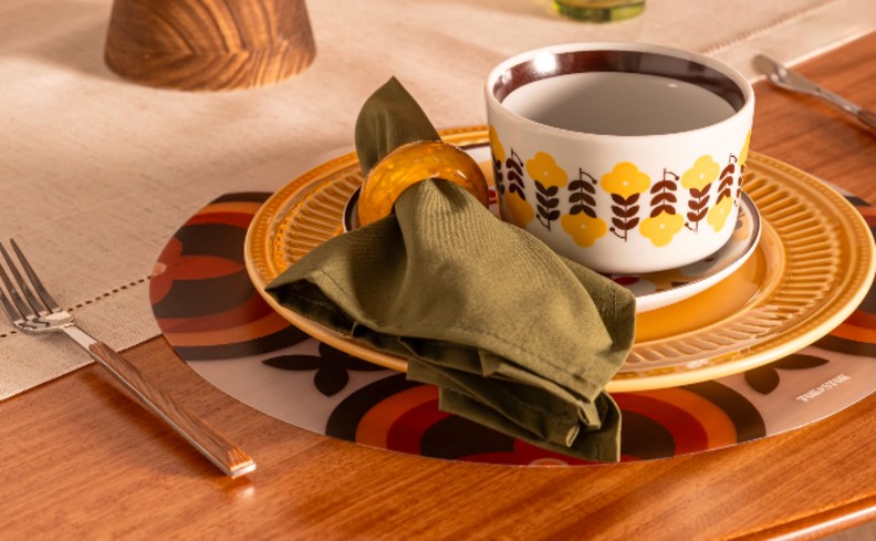 guardanapo de tecido mesa posta para café da tarde com prato, caneca, guardanapo de tecido com argola de guardanapo amarelo, além de sousplat e talheres.