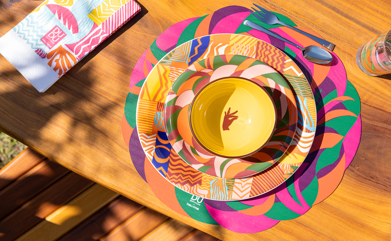 mesa posta para café da tarde com prato, tigela, sousplat, guardanapo e taça com muitas cores vibrantes.