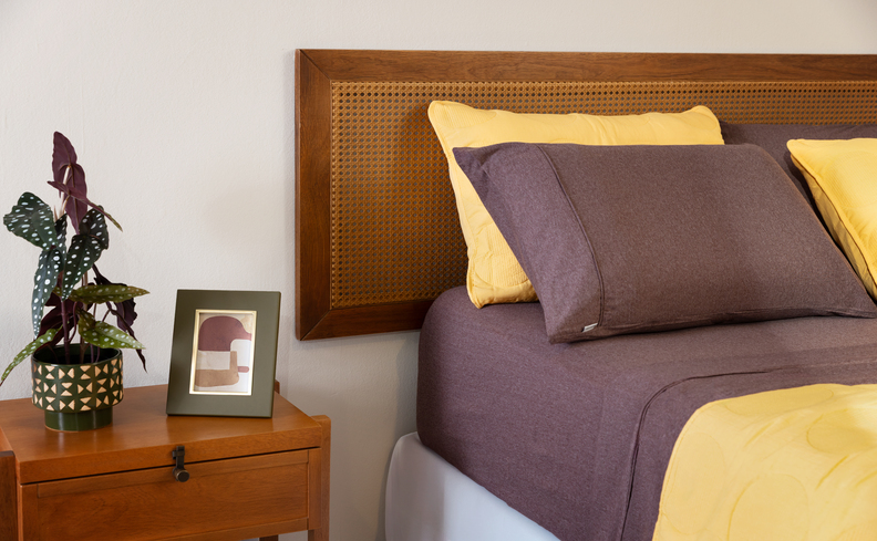 altura de cabeceira de cama de casal em um modelo de palhinha, cama posta em tons de amarelo e marrom, além de mesa de cabeceira com vaso de flores e um porta-retrato.