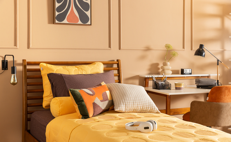 cabeceira de cama solteiro em madeira, cama posta com tons de amarelo e marrom, além de escrivaninha ao lado da cama, arandela e quadro decorativo na parede.